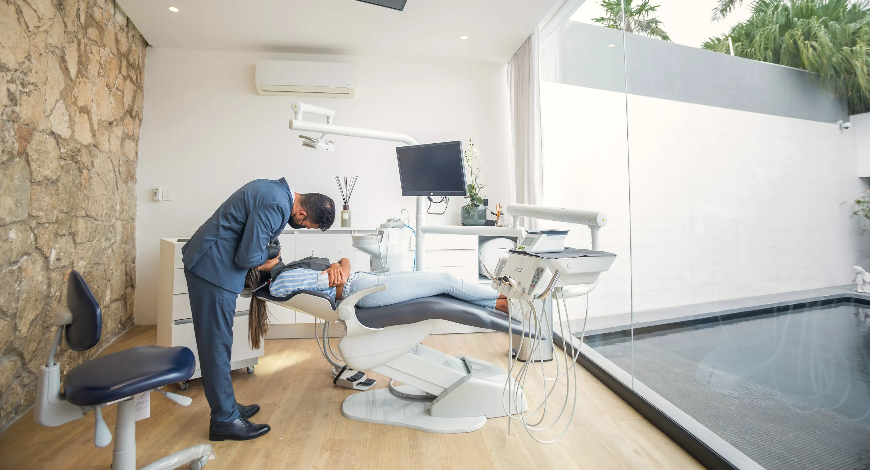 A treatment room in daylight: a stone wall, a wooden floor and a glass wall onto a courtyard pool, a dentist at the chair.